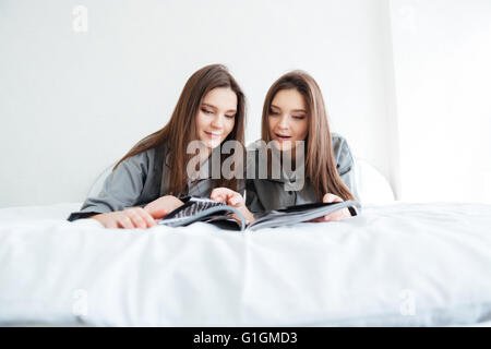 Zwei glückliche schöne junge Schwestern Zwillinge am Bett liegen und lesen Magazin Stockfoto