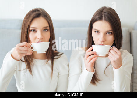 Zwei süße schöne junge Schwestern Zwillinge auf Sofa sitzen und Kaffee trinken Stockfoto