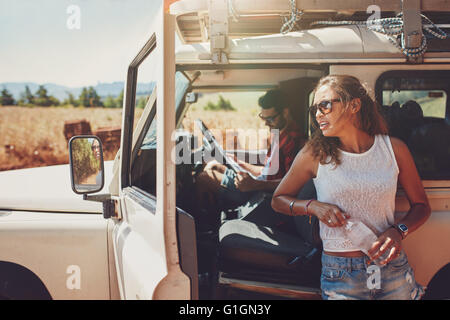 Junge Frau, die außerhalb des Autos hält eine Flasche Wasser mit Mann sitzt auf dem Fahrersitz Blick auf eine der Road Map. Paar o Stockfoto