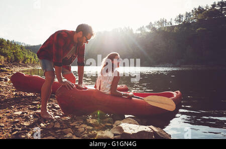 Junger Mann schiebt eine Kanu in das Wasser, während eine Frau sitzt auf dem Kanu. Paar für Kajak in See an einem Sommertag geht. Stockfoto