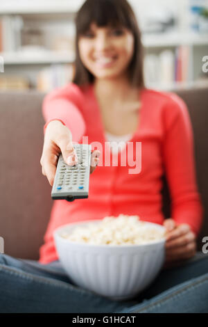 Junge, lächelnde Frau zu Hause auf der Couch sitzen und Fernsehen, sie hält eine Fernbedienung und Essen popcorn Stockfoto