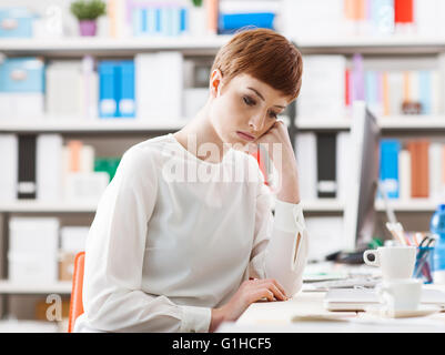 Sad gelangweilt Frau am Schreibtisch sitzen, sie ist depressiv, Blick nach unten und stützte sich auf ihre hand Stockfoto