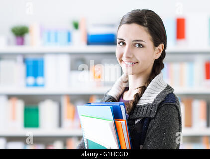 Lächelnde junge Studentin posiert in der Schulbibliothek, sie hält Notizbücher und Blick in die Kamera Stockfoto