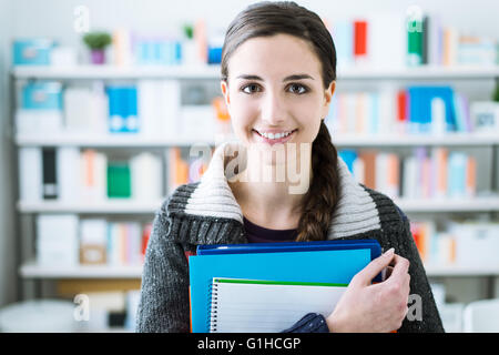 Lächelnde junge Studentin posiert in der Schulbibliothek, sie hält Notizbücher und Blick in die Kamera Stockfoto