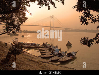 Land Angelboote/Fischerboote am Ufer des Hooghly River während des Sonnenuntergangs mit Second Hooghly Bridge im Hintergrund, Kolkata, Indien Stockfoto