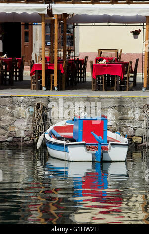 Fisch Taverne rote Tischdecke bedeckt Tische mit Blick auf die Promenade Kai & ein Boot im Vordergrund. Lemnos Insel, Griechenland Stockfoto