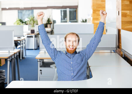 Happy aufgeregt bärtigen Geschäftsmann mit erhobenen Händen sitzen und feiern Erfolg im Büro Stockfoto