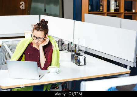 Traurig süße junge Geschäftsfrau in Decke arbeiten mit Laptop und Kältegefühl im Büro Stockfoto