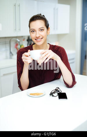 Fröhliche charmante junge Frau trinken Tee und Essen Cookie auf die Küche Stockfoto