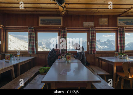 Junges Paar beim Abendessen im Speisesaal Tölzer Hütte mit dem Panorama der schneebedeckten Karwendelgebirge bei Sonnenuntergang, Tirol, Österreich Stockfoto