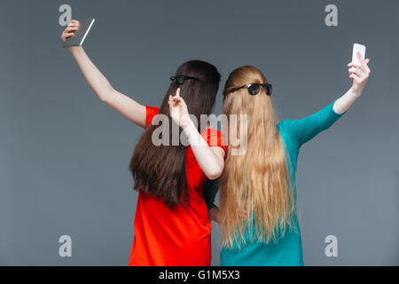 Zwei lustige junge Frauen Gesicht mit langen Haaren und nehmen Selfie mit Tablet und Smartphone überdeckt grauen Hintergrund Stockfoto