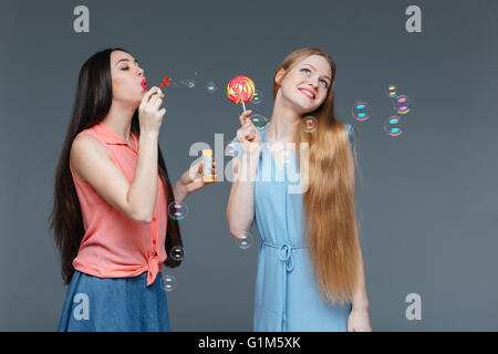 Zwei fröhliche schöne junge Frauen Essen bunte Lutscher und Seifenblasen über grauen Hintergrund Stockfoto