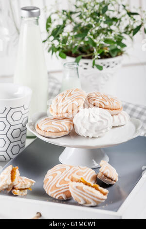 Cookies auf weißen und grauen Platte mit Tassen Kaffee und Flaschen Milch auf weißen Fensterläden Hintergrund mit grünen und weißen Blume Stockfoto