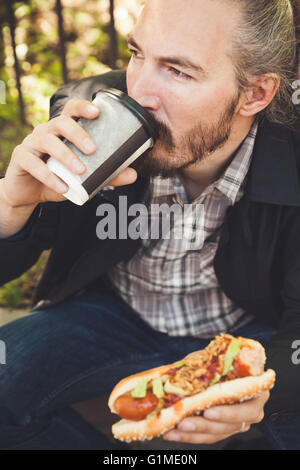Bärtiger asiatischen Mann Mittagessen mit Hot Dog und Kaffee im Sommerpark, Outdoor-Portrait mit selektiven Fokus Stockfoto