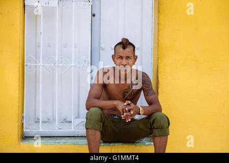 Junger Mann mit Tattoo sitzt vor der Haustür des gelben Hauses in Santiago De Cuba, Kuba. Stockfoto