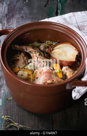 Braune Keramiktopf mit Geschmortes Kaninchen mit Bouillon, Gemüse und Kräutern, serviert mit Küchentuch über alten Holztisch. Rusti Stockfoto