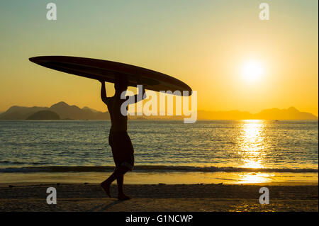 Silhouette des brasilianischen Mann trägt ein Longboard Surfbrett auf dem Kopf Copacabana Strand entlang vor einem goldenen Sonnenaufgang Stockfoto