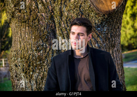 Hübscher junger Mann lehnte sich gegen Baum, wegschauen, eine Seite an einem sonnigen Tag Stockfoto