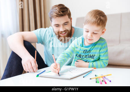 Glückliche kleine Sohn und Papa sitzen und zeichnen mit bunten Markierungen am Tisch Stockfoto