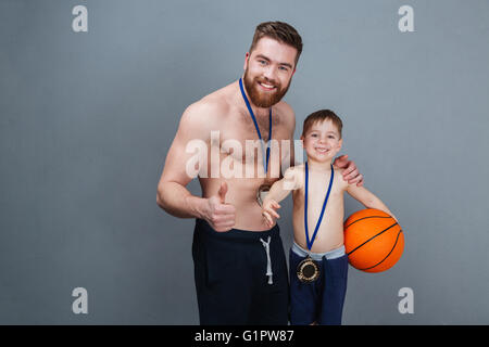 Lächelnd Vater und seinen kleinen Sohn mit goldenen Medaillen mit Basketball Ball über grauen Hintergrund Stockfoto