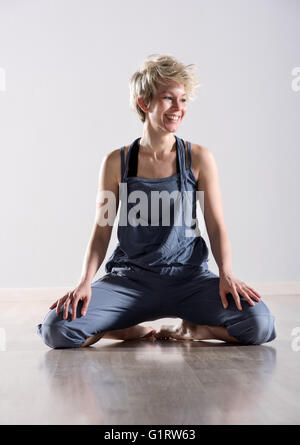 Glücklich lachende einzelne junge Frau im blauen Outfit im sitzen mit den Zehen auf Parkett über grauen Hintergrund