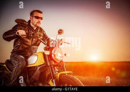 Biker Mann trägt eine Lederjacke und eine Sonnenbrille sitzt auf seinem Motorrad, Blick auf den Sonnenuntergang, Rennen auf der Straße. Filter-ap Stockfoto