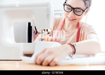 Lächelnde hübsche junge Frau Schneiderin näht an der Nähmaschine in Werkstatt Stockfoto