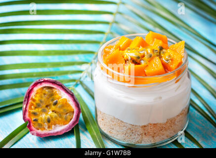 Käsekuchen im Glas von tropischen Früchten: Mango und Passionsfrucht, Palmzweig auf blauem Untergrund aus Holz Stockfoto