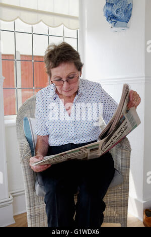Ältere Frau zog sich Reife Lesung Lokalzeitung im Korbstuhl zu Hause sitzen Stockfoto
