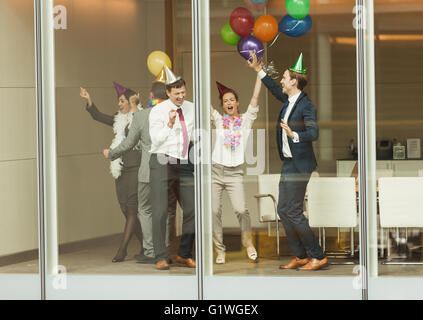 Business-Leute tragen Partyhüte und Tanz mit Luftballons an Konferenz Zimmer Fenster Stockfoto