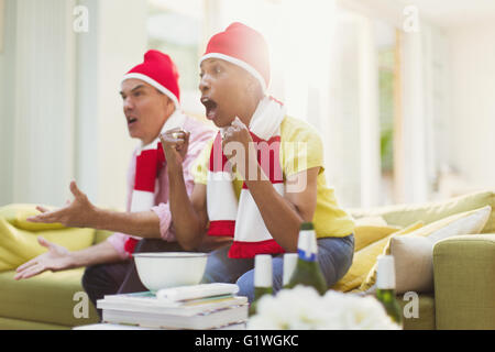 Älteres Paar tragen passende Hüte und beobachten TV-Sport-Event im Wohnzimmer jubeln Stockfoto