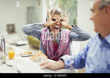 Spielerische Reife Frau gestikulieren Finger Gläser am Frühstückstisch Stockfoto