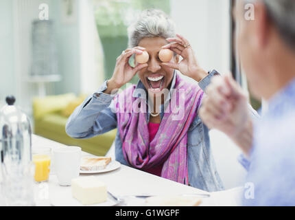 Spielerische Reife Frau für Augen mit Eiern am Frühstückstisch Stockfoto