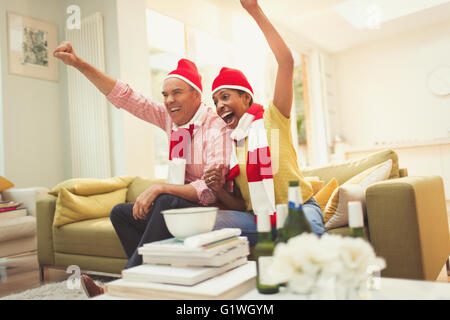 Begeisterte älteres Paar in passenden Mützen und Schals, jubeln, vor dem Fernseher Sportveranstaltung Stockfoto