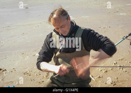 Fischer in Watvögel kniete auf dem nassen Sand hält eine Angelrute und wickeln die Linie auf der Spule mit einem schnellen Handgriff Stockfoto