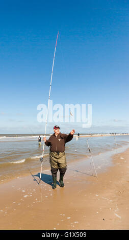 Fischer mit Angelrute und zwei gefangenen Fische auf der Linie stehend am Nordseestrand gekleidet in Watvögel, Pullover und Mütze Stockfoto