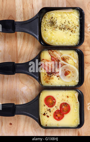 Foto von einem drei Raclette-Tabletts mit geschmolzenem Käse, Kräuter, Kirschtomaten, Speck und Zwiebeln. Stockfoto