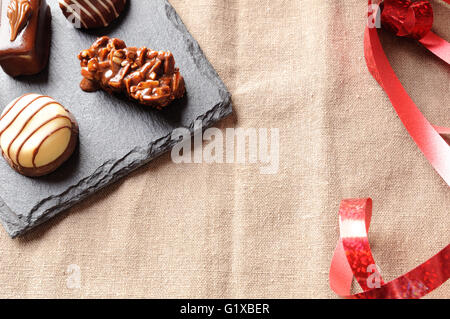 Sortierten Bonbons schwarze und weiße Schokolade mit Nüssen und rotes Band auf einer Schiefertafel Platte in einer Tabelle mit braun Tischdecke Stoff. Stockfoto