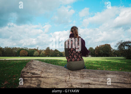 Eine junge Frau sitzt auf einem großen umgestürzten Baum im Park an einem sonnigen Herbsttag Stockfoto