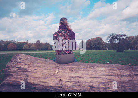 Eine junge Frau sitzt auf einem großen umgestürzten Baum im Park an einem sonnigen Herbsttag Stockfoto