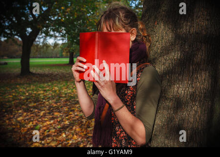 Eine junge Frau befindet sich im Park an einem sonnigen Herbsttag und versteckt sich hinter einem Buch Stockfoto