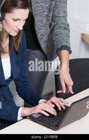 Eine junge Geschäftsfrau mit Kopfhörer ist bei einem Notebook eingeben. Eine andere Frau zeigt auf dem Display. Stockfoto