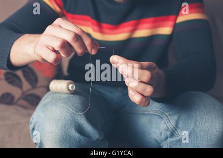 Ein junger Mann sitzt auf dem Sofa und ist eine Nadel einfädeln Stockfoto