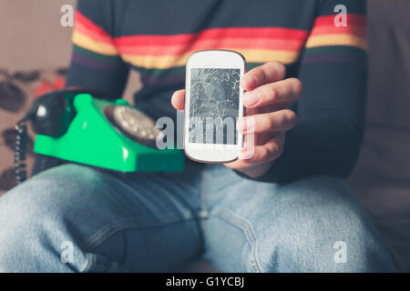 Ein junger Mann sitzt auf einem Sofa mit einem gebrochenen Smartphone und einem alten Telefon mit Wählscheibe Stockfoto