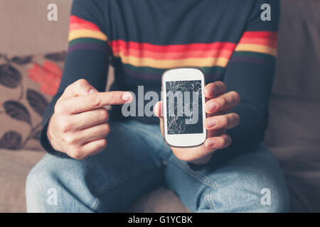 Ein junger Mann sitzt auf dem Sofa und ist auf eine gebrochene Smartphone mit einem gebrochenen Bildschirm zeigen Stockfoto