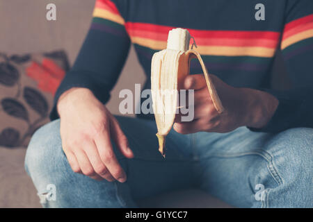 Ein junger Mann sitzt auf einem Sofa und hält eine Banane Stockfoto