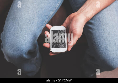 Ein junger Mann sitzt mit einem gebrochenen Smartphone Stockfoto