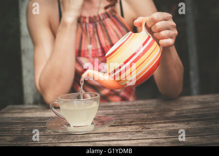 Eine junge Frau sitzt an einem Tisch und gießt sich eine Tasse Tee Stockfoto