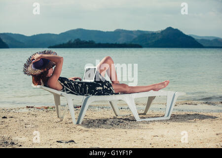Eine junge Frau sitzt auf einer Liege an einem tropischen Strand und arbeitet an einem laptop Stockfoto