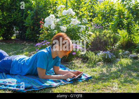 Frau mit Brille und lässige Kleidung arbeiten mit Tablet liegend im Freien im blühenden Garten. Natürliches Tageslicht, echte peopl Stockfoto